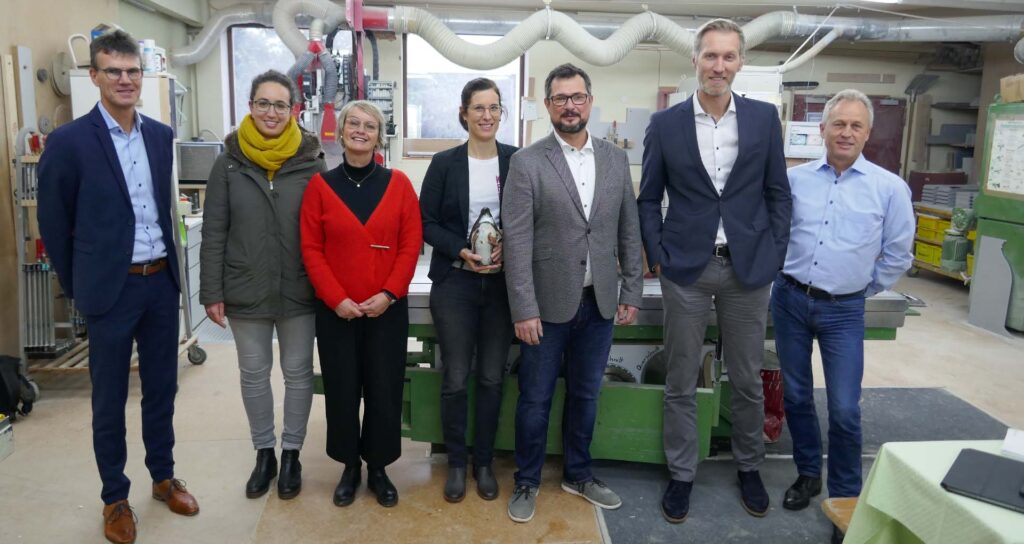 Bild zeigt alle Beteiligten v.l.n.r.: Dippold, Saft, Seeger, Protze, Protze, Terzenbach, Mevenkamp in deer Werkstatt der Schreinerei Protze. Sie stehen in einer Reihe nebeneinander und lächeln in die Kamera. Daniela Protze hält den Pinguin-Award in den Händen.