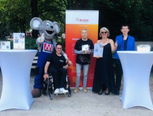 Vier Personen posieren zusammen mit dem Maskottchen der Firma Mauss Bau für ein Foto. Das Maskottchen ist eine große graue Maus.