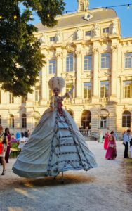 Eine Frau in einem ausladenden barocken Kleid läuft auf Stelzen durch einen geschmückten Park. Sie trägt eine weiße Perücke und ist auffällig geschminkt.