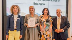 Vier Personen stehen nebeneinander. Die zweite Person von links, eine Frau im grauen Kleid, hält einen Preis in den Händen.