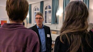 Thomas Wedel im Gespräch. Im Vordergrund sind unscharf die Hinterköpfe seiner Gesprächspartnerinnen zu sehen.