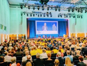 Ein voll besetzter Veranstaltungssaal. Der Saal ist in grünliches Licht getaucht. Die Bühne ist weit entfernt, über der Bühne ist ein großer Monitor.