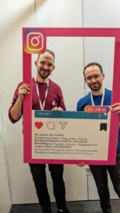 Zwei Personen schauen durch einen pinken Rahmen. Auf dem Rahmen steht: Wir passen alle ins Bild.